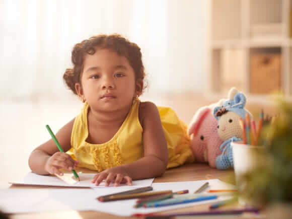 menina com blusa amarela segurando um lápis e desenhando em um caderno