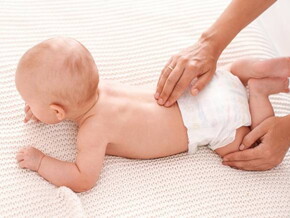 bebê deitado de bruços e uma mão segundo sua perna e outra massageando sua lombar