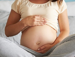 Gestante sentada na cama com as pernas cruzadas enquanto acaricia a barriga com as duas mãos.