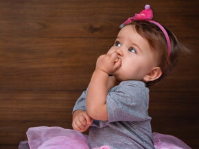 Menina de aproximadamente um ano usando vestido rosa e com a mão na boca.