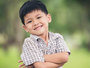 Menino asiático usando camisa xadrez sorrindo com os braços cruzados.