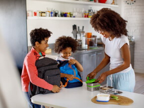 duas crianças com suas mochilas aguardam mãe fechar suas lancheiras