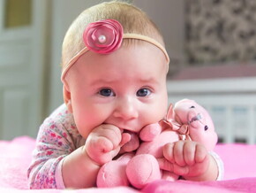 Bebê recém nascida chamada Liz deitada na cama com um ursinho de pelúcia na boca.