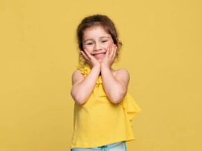 Uma menina sorridente de aproximadamente 3 anos, usando uma blusa amarela, de costas para um fundo amarelo.