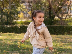 Uma menina de aproximadamente 4 anos brincando ao ar livre em um parque, com um casaco bege e tranças.