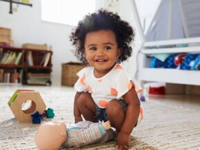 Bebê sorridente de cabelos crespos agachada sobre um tapete, brincando com uma boneca em um ambiente infantil.