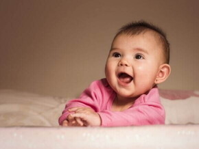 Bebê sorridente deitada de bruços na cama usando uma roupa cor de rosa.