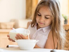 Menina sentada na mesa de jantar despejando leite em um tigela de cereais