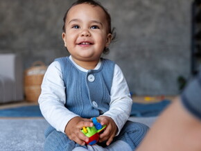 Bebê de aproximadamente 1 ano sentado no tapete da sala segurando brinquedos.