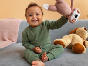 Menino usando macacão verde sentado no sofá segurando ursinho de pelúcia e sorrindo