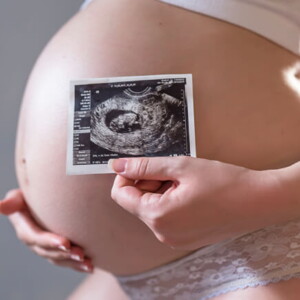 barriga grávida com uma das mãos posicionadas na barriga e a outra segurando a imagem de uma ultrassonografia