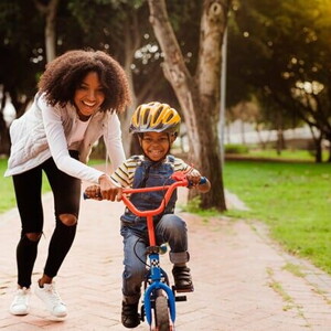 Foto de um menino sorrindo, em uma bicicleta sem rodinhas e ao lado uma mulher adulta também sorrindo segurando em um dos lados do guidão da bicicleta.