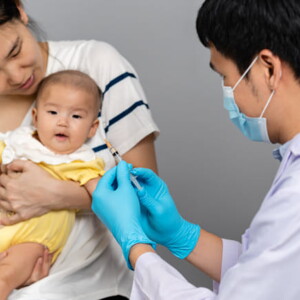 imagem de um bebê no colo de uma mulher adulta e um homem segurando uma seringa, vestindo roupas de profissionais da saúde na cor branca