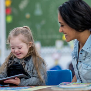 Criança com Síndrome de Down segurando um tablet ao lado da professora na escola