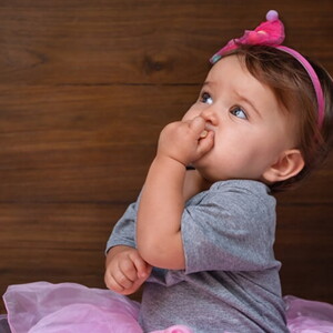 Menina de aproximadamente um ano usando vestido rosa e com a mão na boca.