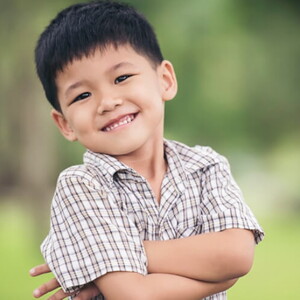 Menino asiático usando camisa xadrez sorrindo com os braços cruzados.