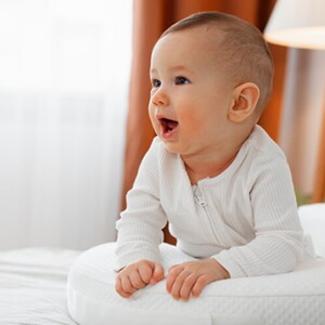 Bebê usando macacão branco deitado de barriga para baixo em cima da cama.