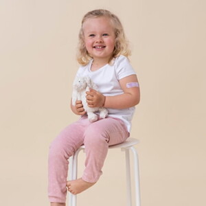Menina loira de aproximadamente quatro anos sentada em uma cadeira segurando um ursinho de pelúcia.