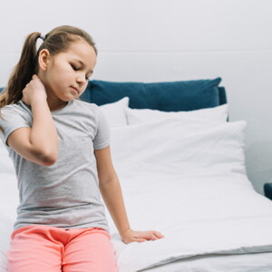 imagem de uma menina sentada na beira de uma cama, com uma das mãos no pescoço e com olhos fechados