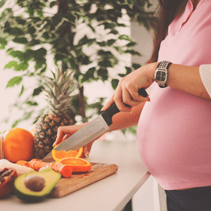 Pregnant woman slicing an orange by a kitchen counter full of fruit.