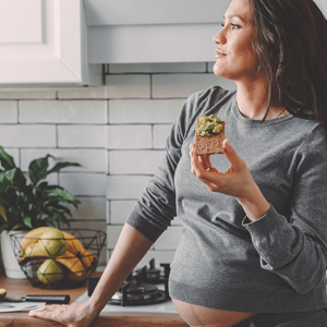 Dreamy looking pregnant woman, standin on her kitchen eating an avocado toast