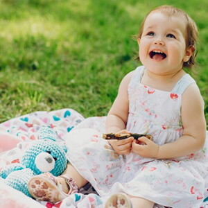 Menina de aproximadamente um ano usando vestido florido e sentada na grama de um parque.