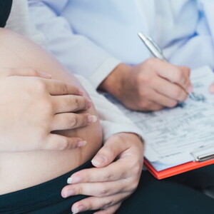 Médico com uma prancheta nas mãos fazendo anotações enquanto uma mulher grávida está sentada na cama