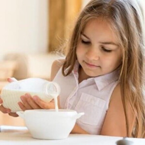 Menina sentada na mesa de jantar despejando leite em um tigela de cereais