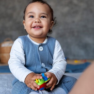 Bebê de aproximadamente 1 ano sentado no tapete da sala segurando brinquedos.