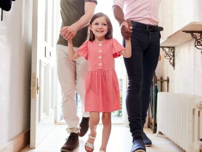 Uma menina com aspecto de felicidade, andando de mãos dadas com dois homens em um ambiente doméstico.