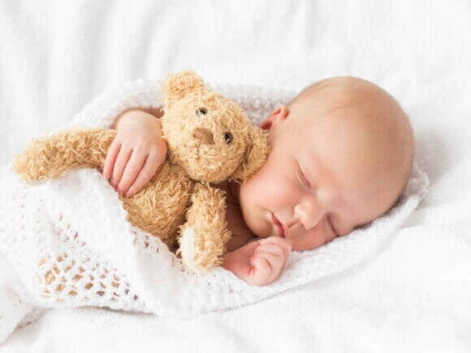 Bebê dormindo tranquilamente enrolado em uma manta branca e abraçado em um urso pequeno de pelúcia.