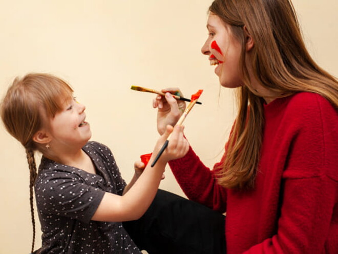 mulher com cabelos longos e loiros, vestindo suéter vermelho, segurando um pincel de tinta, está sorrindo enquanto olha para uma menina com cabelos loiros com tranças e que também segura um pincel de tinta e sorri.