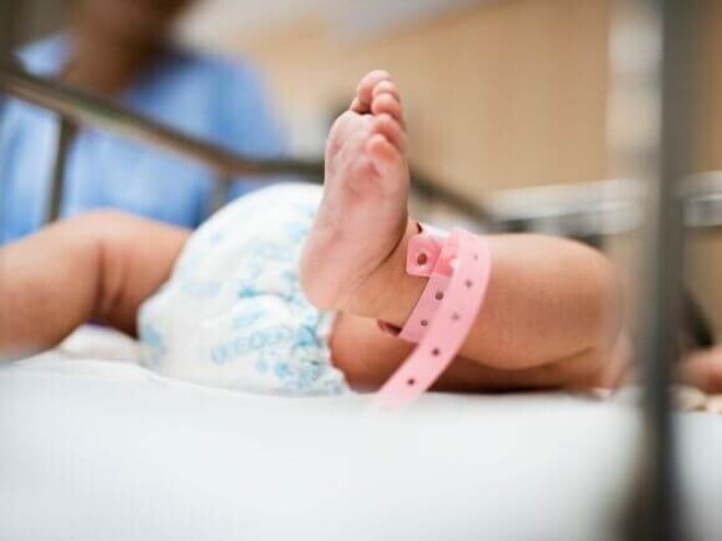 Close de um bebê recém-nascido deitado em um berço hospitalar, com uma pulseira rosa de identificação no tornozelo, simbolizando cuidados neonatais e saúde infantil.