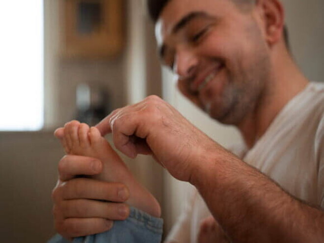 Um homem sorrindo e segurando o pé de uma criança fazendo um movimento de cócegas com o dedo.