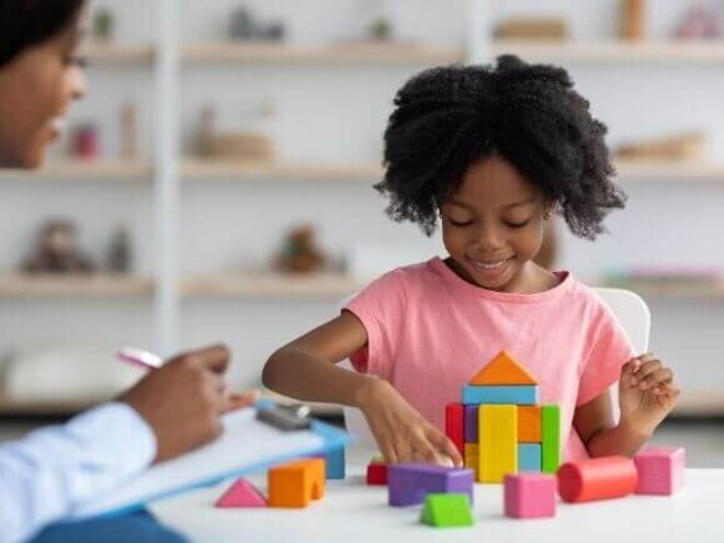 Uma menina sorrindo e organizando blocos coloridos, ao lado de uma mulher sentada com uma ficha na mão.