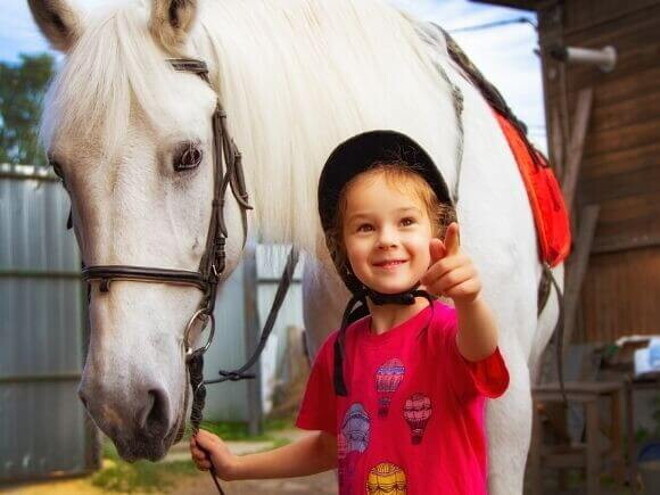 Menina sorrindo e apontando com o dedo, enquanto segura a rédea de um cavalo