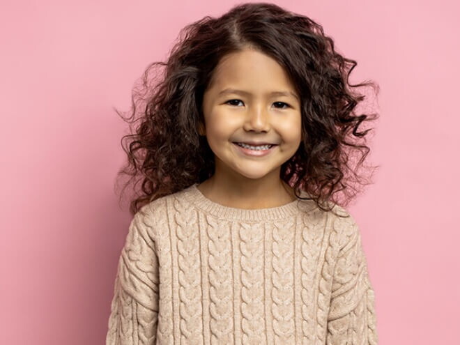 Menina de cabelo ondulado sorrindo em frente a uma parede cor de rosa.