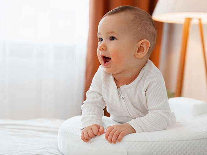 Bebê usando macacão branco deitado de barriga para baixo em cima da cama.