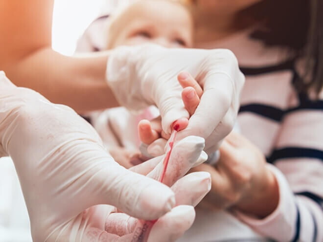 Mãe segurando bebê enquanto enfermeira colhe sangue para exame.