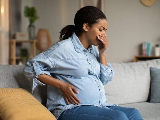 Mulher grávida sentada em um sofá, com uma mão na barriga e a outra mão na boca.