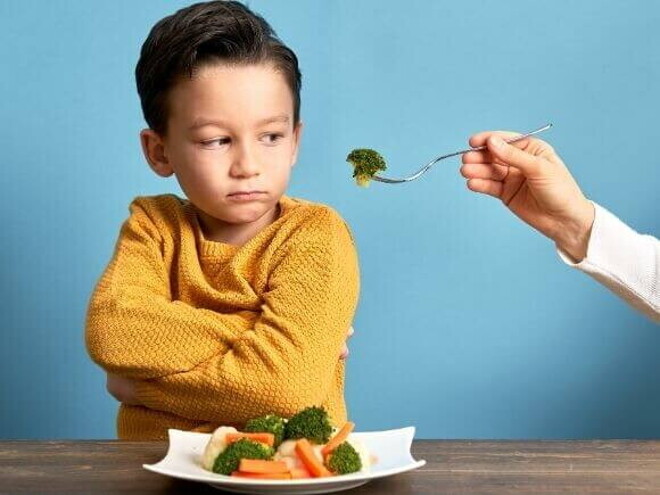 Uma mão estendendo um garfo com brócolis para um menino, que olha de forma irritada para o alimento.