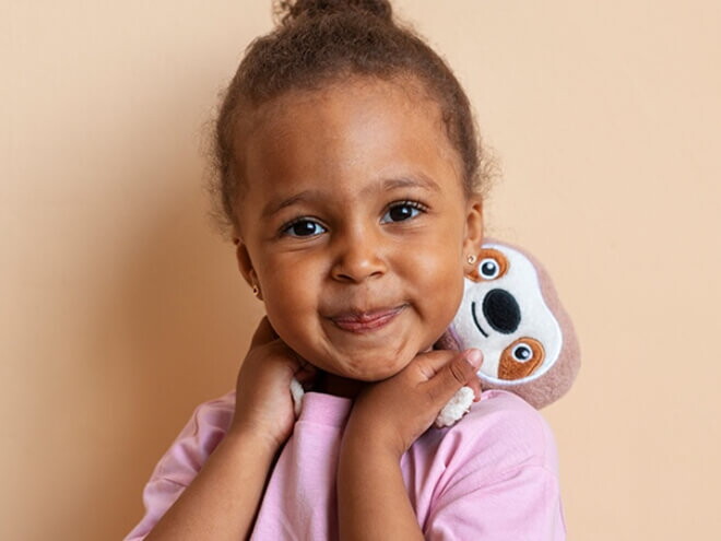 Menina sorrindo vestindo blusa lilás e segurando um bicho preguiça de pelúcia