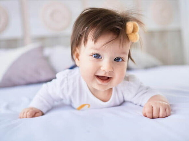 Bebê sorridente deitada de bruços na cama, usando roupa branca e um laço no cabelo