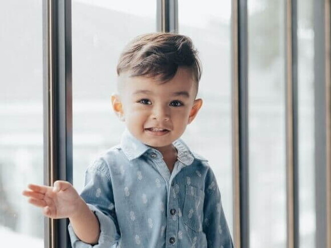 Menino sorrindo com uma camisa azul, com uma porta de vidro ao fundo.