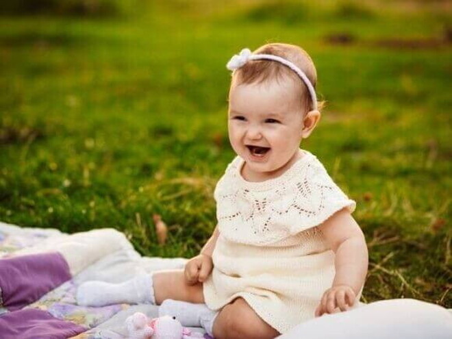 Bebê sorridente usando um vestido claro e uma tiara com flores, sentada em um cobertor sobre a grama.