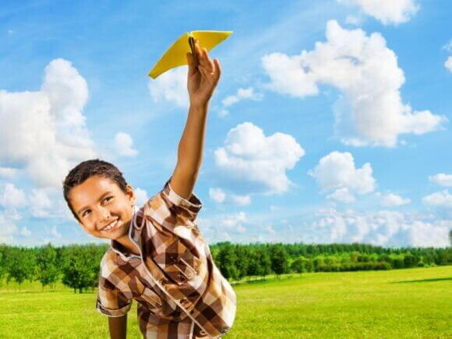 Menino sorridente com aproximadamente 07 anos brincando em um campo verde com um aviãozinho de brinquedo.