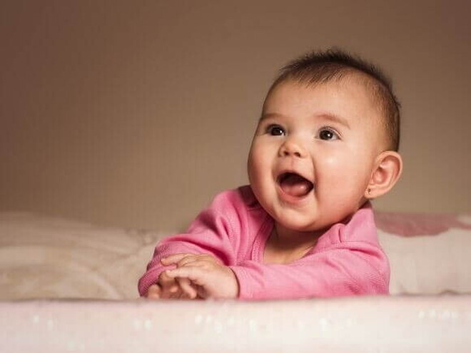 Bebê sorridente deitada de bruços na cama usando uma roupa cor de rosa.