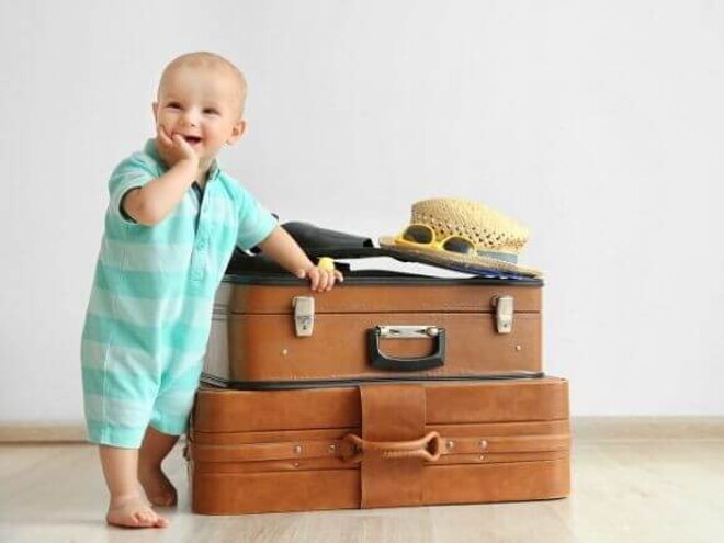 Bebê sorrindo, com a mão na boca, apoiado em malas de viagem.