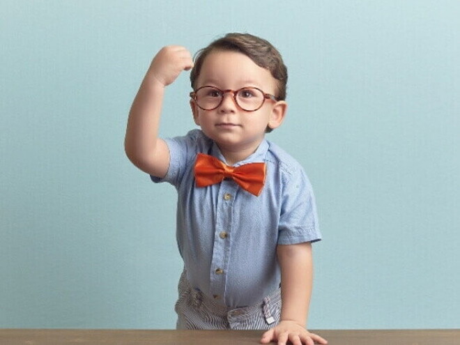 Menino com óculos, gravata vermelha e camisa azul, está apoiado sobre uma mesa, com a mão direita levantada em punho, sob um fundo azul.