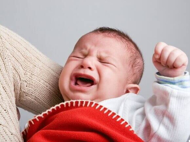 Um bebê chorando intensamente, enrolado em um cobertor vermelho, com o rosto virado para cima, demonstrando grande desconforto.